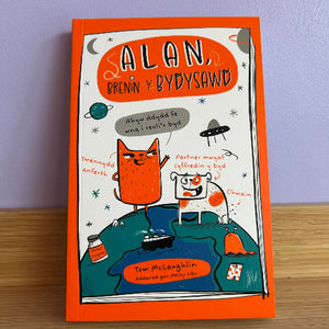 Book cover of 'Alan, Brenin y Bydysawd' with an orange border on a wooden surface.