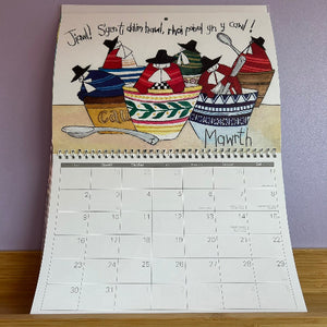 Calendar with Welsh language text and traditional food illustration on a wooden surface.