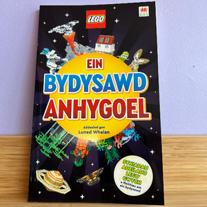 LEGO book titled 'Ein Bydysawd Anhygoel' on a wooden surface with a purple wall background