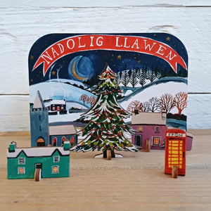 Christmas decoration with a snowy village scene, tree, and red phone box on a wooden surface.