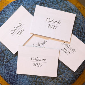 Five white paper cards printed with 'Calendr 2027' arranged on a textured blue fabric surface over a wooden table.
