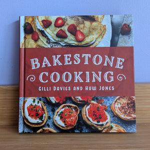 The book 'Bakestone Cooking' by Gilli Davies and Huw Jones stands upright on a light wood shelf against a solid light blue wall. The book cover features a prominent reddish-brown band with white text, flanked by food photography of pancakes and stuffed pastries.