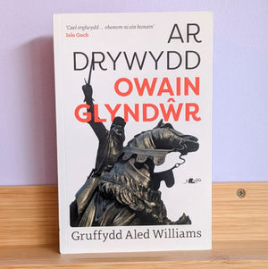A book titled 'Owain Glyndwr' by Gruffydd Aled Williams, featuring a white cover with black and red text and a black-and-white statue illustration, standing on a wooden surface against a light purple wall.