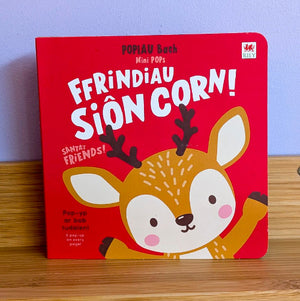 A children's pop-up book titled 'Ffrindiau Siôn Corn!' by Popiau Bach, featuring a cartoon deer illustration on a bright red cover, photographed standing on a wooden surface against a light blue wall.