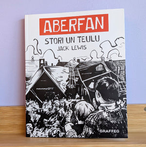 A book titled 'Aberfan: Stori un Teulu' by Jack Lewis, published by Graffeg, standing upright on a wooden shelf against a light purple wall. The cover features a bold red title block with white text on a white background, above a black and white woodcut-style illustration of a crowd looking toward a building.