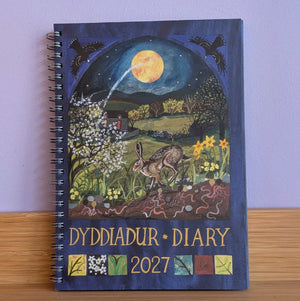 A spiral-bound 2027 Welsh calendar titled 'DYDDIADUR DIARY' featuring a folk-art style illustration of a hare under a full moon on the cover, displayed on a wooden surface against a light purple wall.