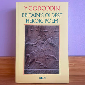 A book titled 'Britain's Oldest Heroic Poem' by Y Goddodin, featuring a cream-colored cover with a central stone relief image of a warrior on horseback, displayed on a wooden surface against a purple wall.