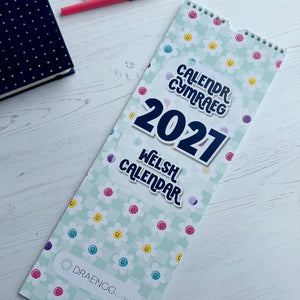 A 2027 Welsh calendar by Draenog Co. lies open on a white wooden tabletop, showing a pastel green floral pattern with colorful polka dots and bold navy text.