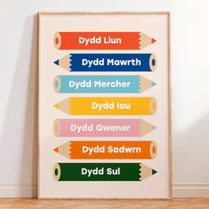 Framed poster with colorful pencil-shaped days of the week labels on a white wall.