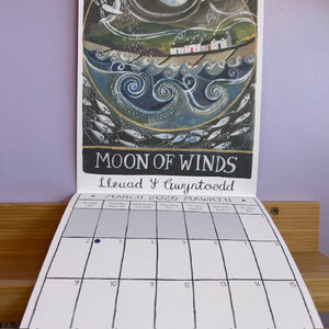 Calendar with a decorative cover titled 'Moon of Winds' on a wooden surface.