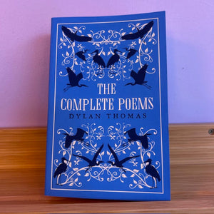 Blue book cover of 'The Complete Poems' by Dylan Thomas with black bird illustrations on a wooden surface.