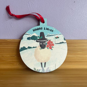 Decorative ornament with a sheep design and 'Nadolig Llawen' text on a wooden surface.