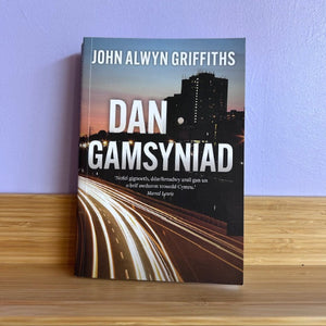 Book titled 'Dan Gamsyniad' by John Alwyn Griffiths on a wooden surface with a blue wall background.