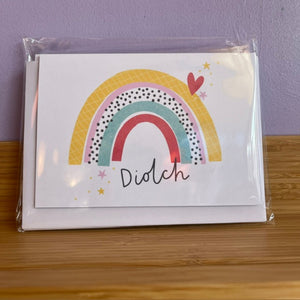 A white greeting card with a colorful rainbow illustration and the word 'Dioclet' written in black script, still sealed in clear plastic packaging, resting on a wooden surface against a lavender wall.
