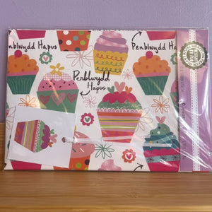 A packaged wrapping paper sheet with a colorful cupcake pattern and the Welsh text 'Penblwydd Hapus' printed on it, wrapped in clear plastic on a wooden surface against a lavender background.
