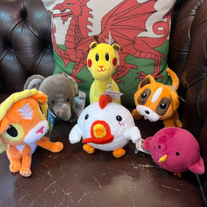 A group of six colorful plush toys arranged on a dark brown leather couch in front of a decorative cushion featuring a red dragon on a green background.
