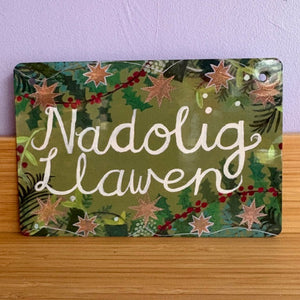 Decorative sign with 'Nadolig Llawen' text on a green background with stars and holly.