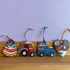 Set of Christmas-themed ornaments including a snow globe, tractor, truck, and heart-shaped ornament on a wooden surface with a purple wall background.