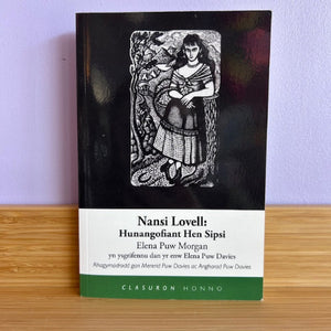 Book titled 'Nansi Lovell: Hunangofiant Hen Sipsi' with a black and white illustration on a wooden surface.