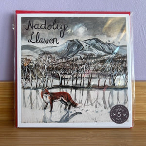 Card with a fox illustration and mountain landscape, featuring the text 'Nadolig Llawen' on a wooden surface.
