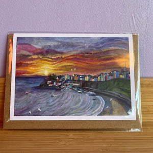 Artistic depiction of a coastal scene with a colorful sunset, framed in clear plastic on a wooden surface.