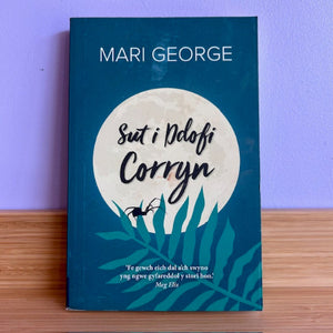 Book titled 'Sut i Dolofi Corryn' by Mari George on a wooden surface with a purple wall background.