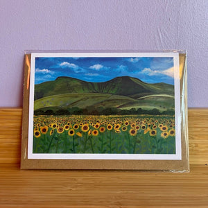 Greeting card with a sunflower field and mountain landscape on a wooden surface.