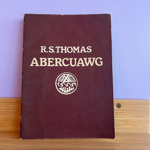 Maroon book titled 'Abercuawg' by R.S. Thomas on a wooden surface with a blue wall background.