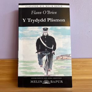 Book cover of 'Y Trydydd Plismon' by Flann O'Brien on a wooden surface with a purple wall background.